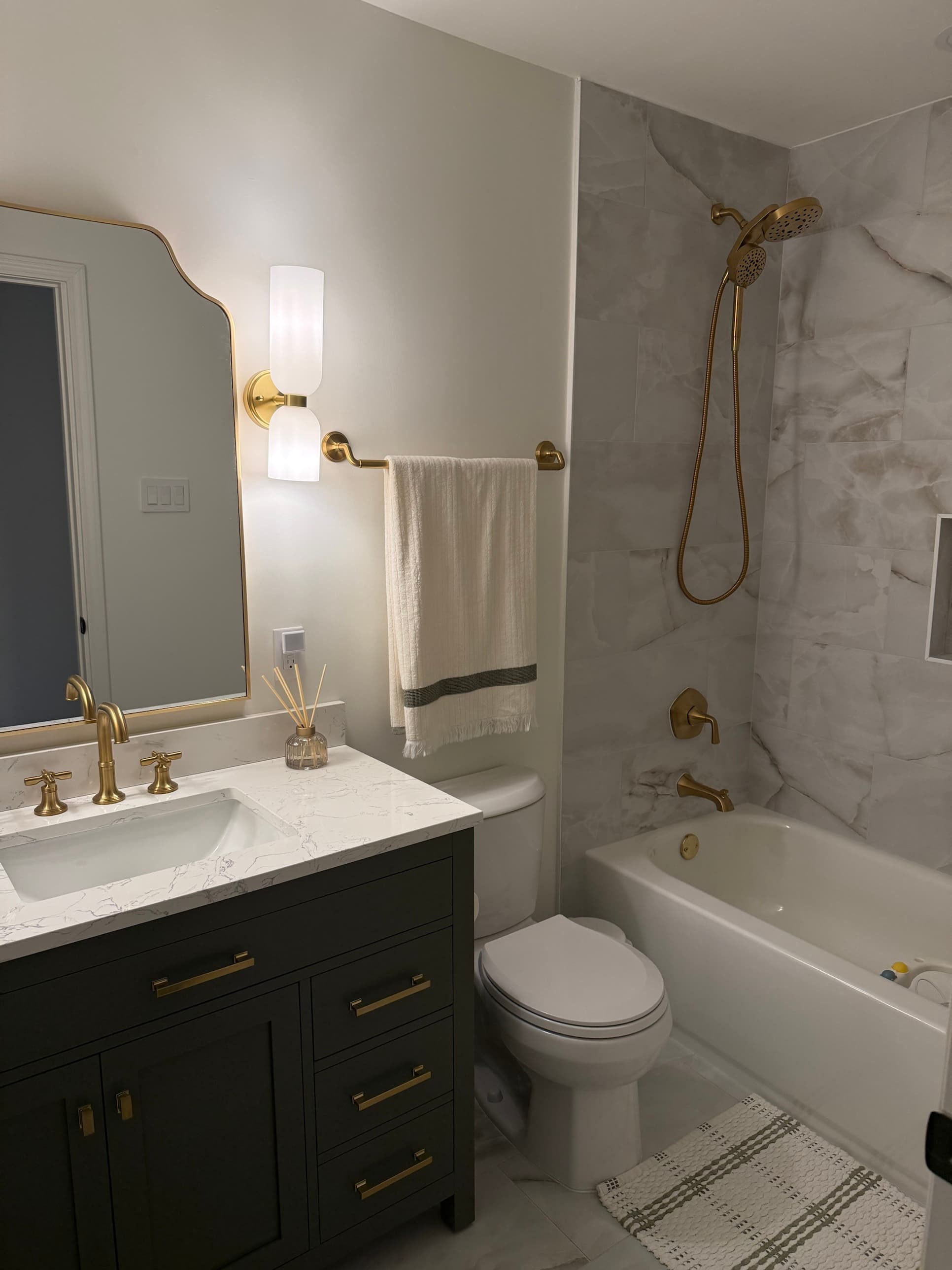 Guest bathroom remodel with dark vanity, gold cross-handle fixtures, marble-look tile shower surround, and quartz countertop in Charlotte, NC